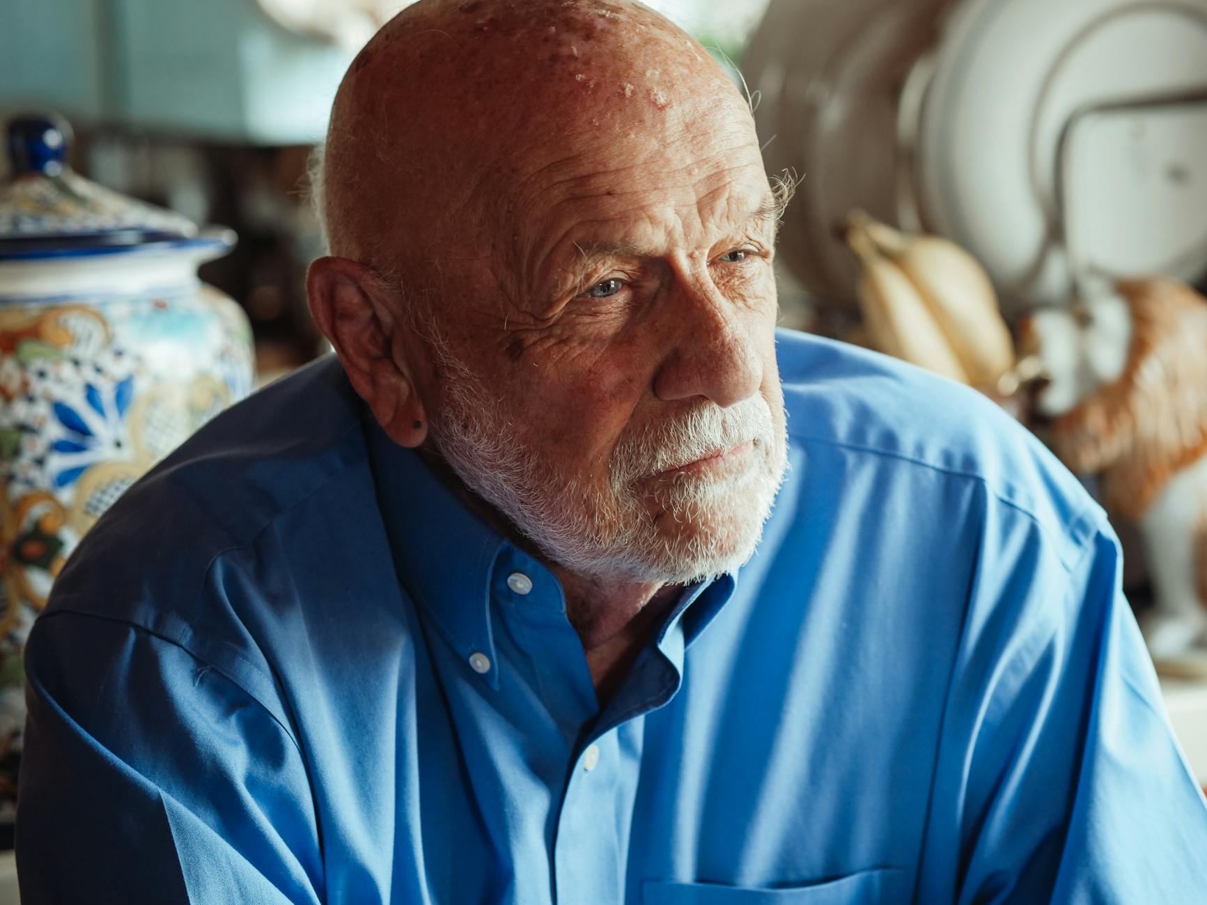 Elderly man with beard in a blue shirt, thoughtfully gazing indoors.