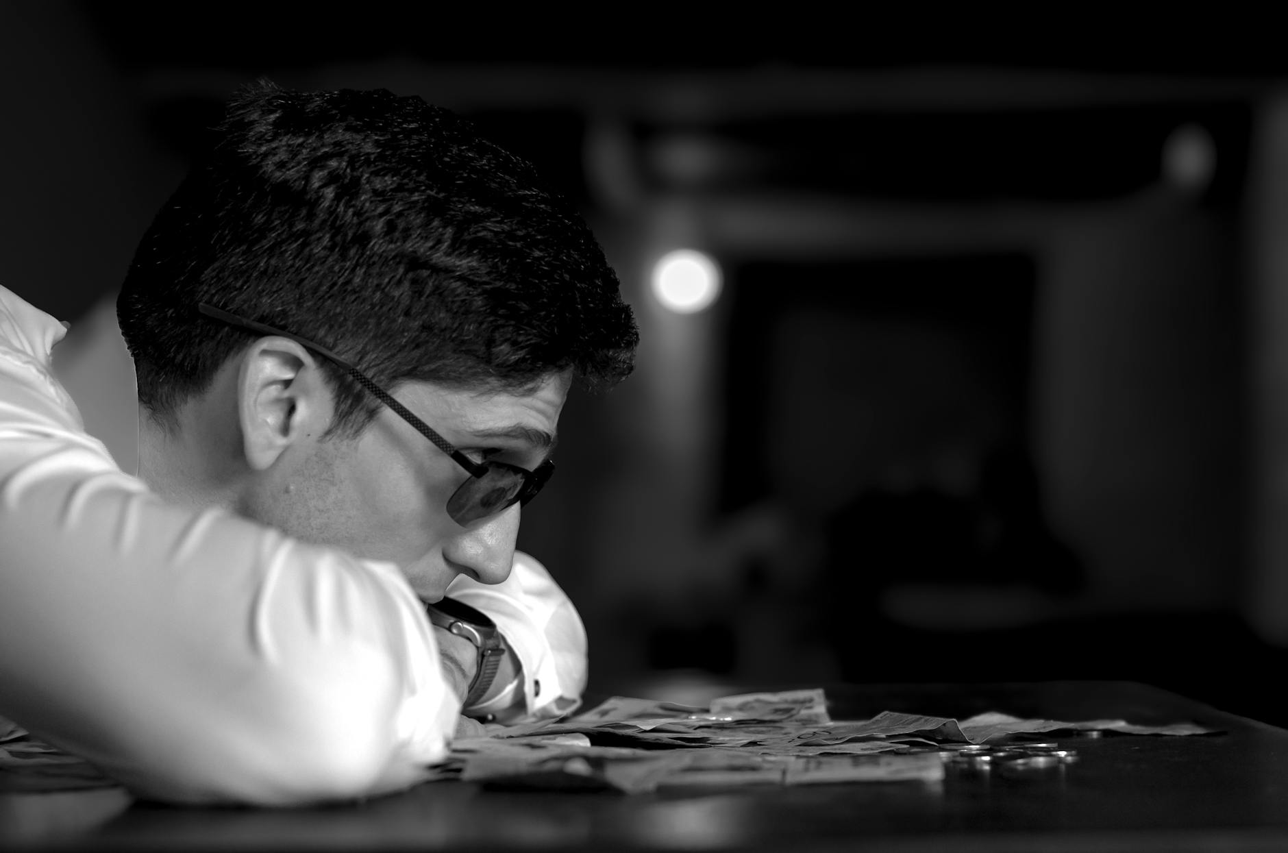 A man in glasses deep in thought, resting on a table with scattered money in a dimly lit room.