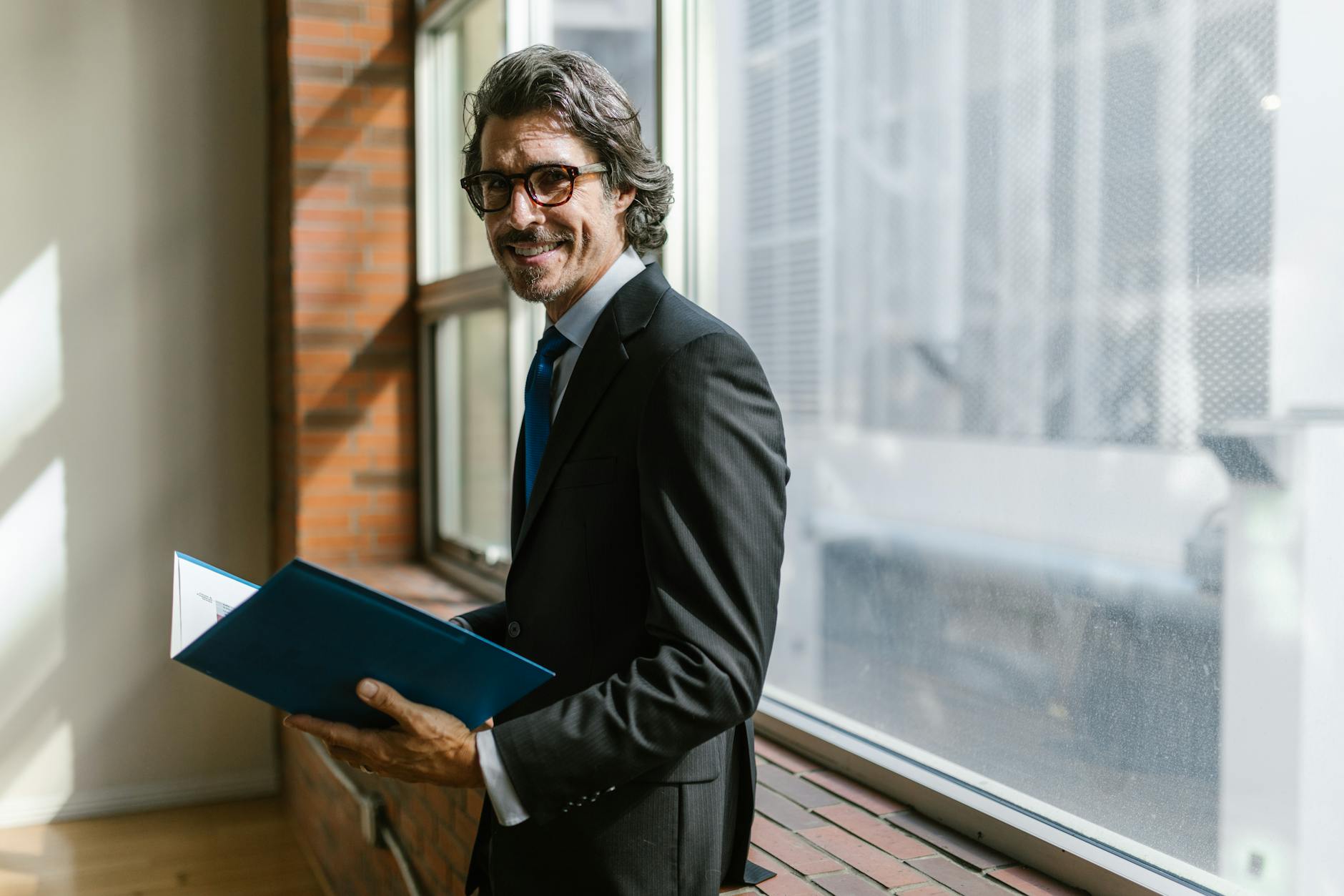 Smiling businessman in a black suit holding a folder by a window indoors.