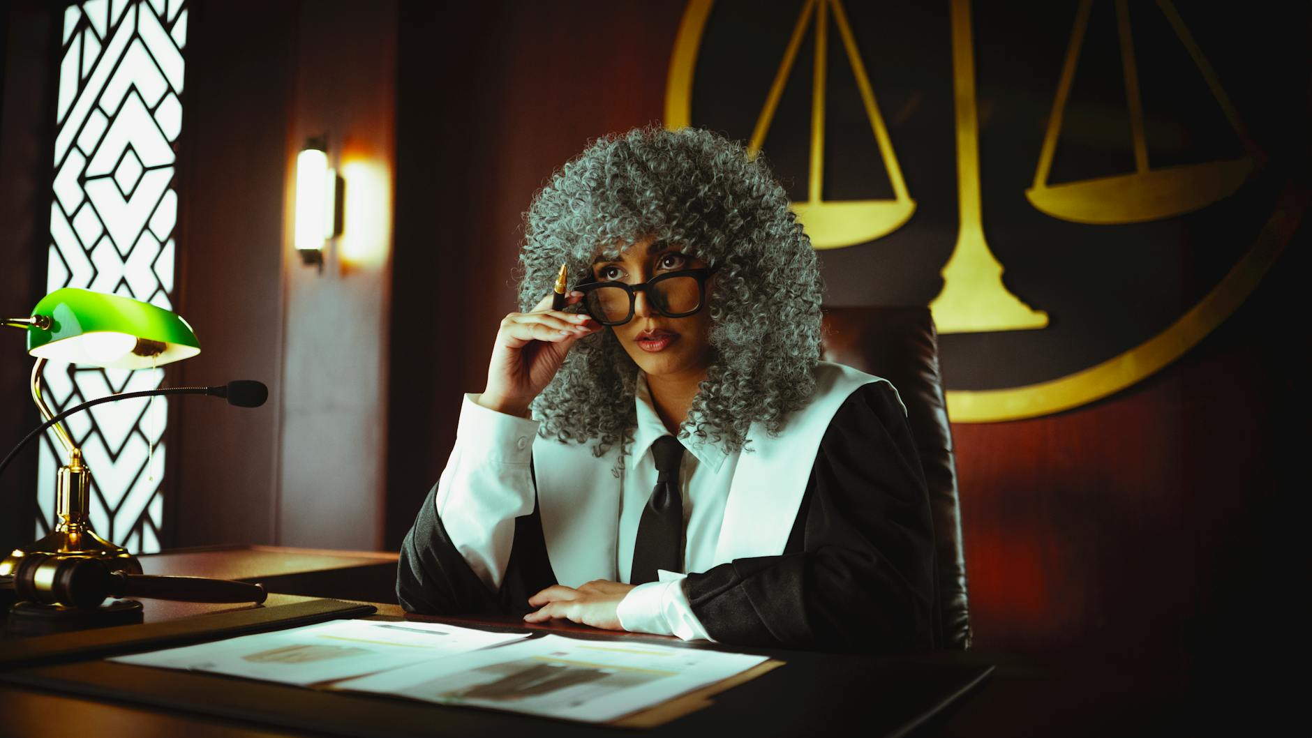 Judge in traditional attire sits at a courtroom desk with a serious expression.