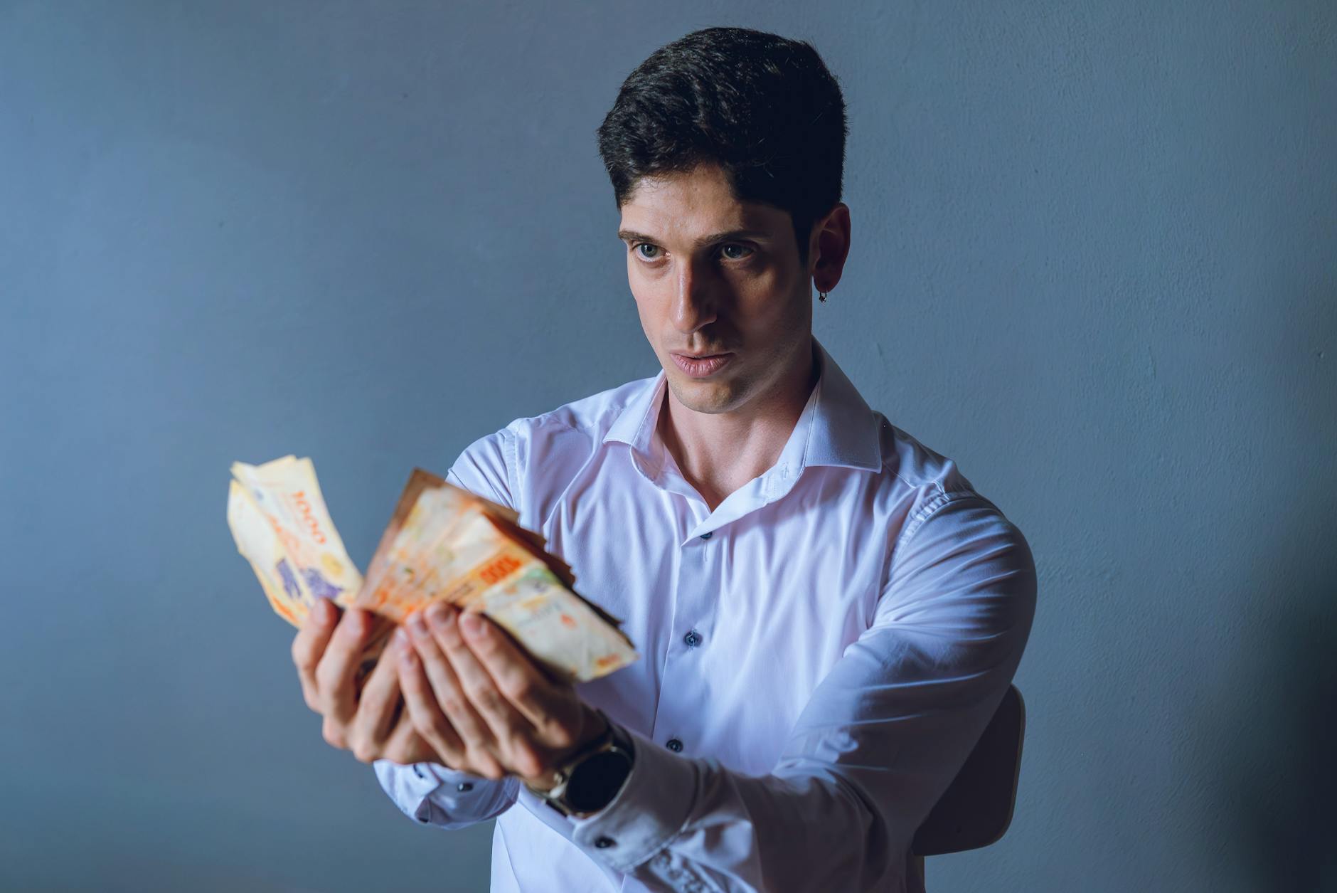A focused adult man counts Argentina pesos indoors, against a neutral backdrop.