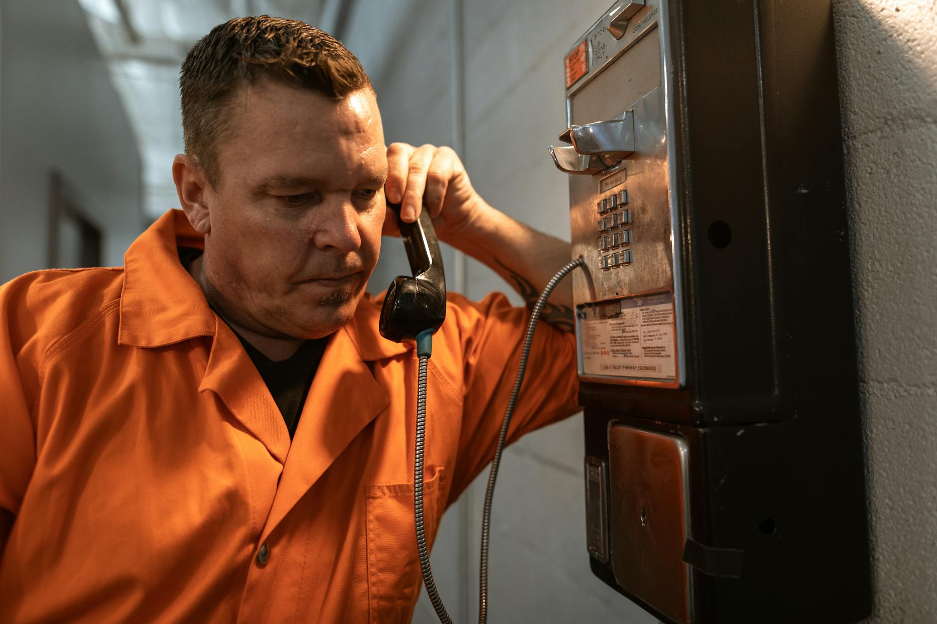 An inmate in an orange jumpsuit makes a phone call inside a prison facility, conveying the prison life atmosphere.
